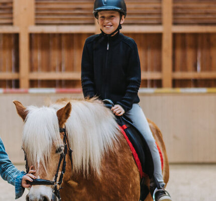 Mädchen reitet auf Pferd, geführt von Reitlehrerin mit Seil Ein Mädchen reitet auf einem Pferd, das von einer Reitlehrerin mit einem Seil geführt wird.